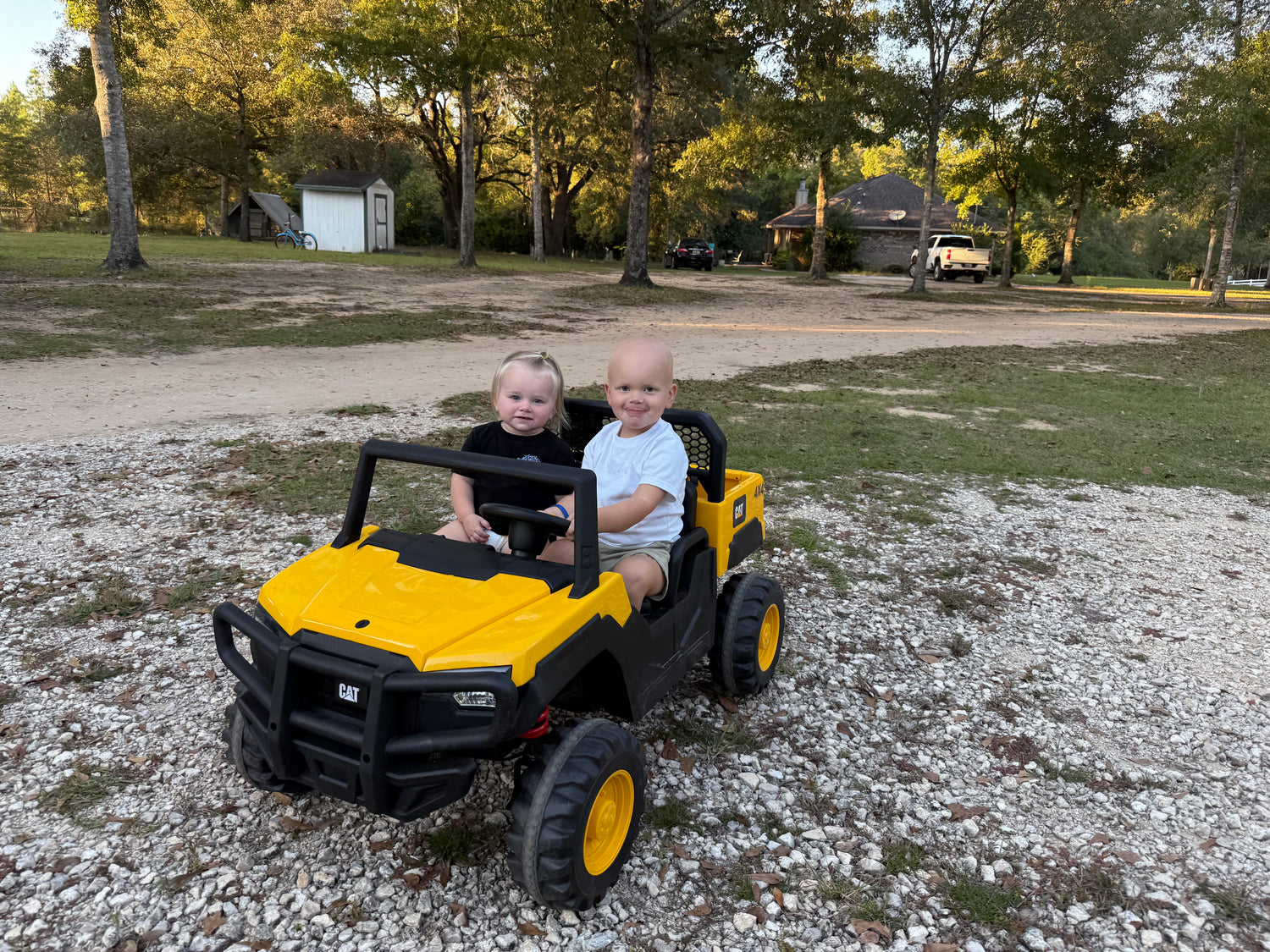 Two children in a yellow and black toy vehicle in a park setting.